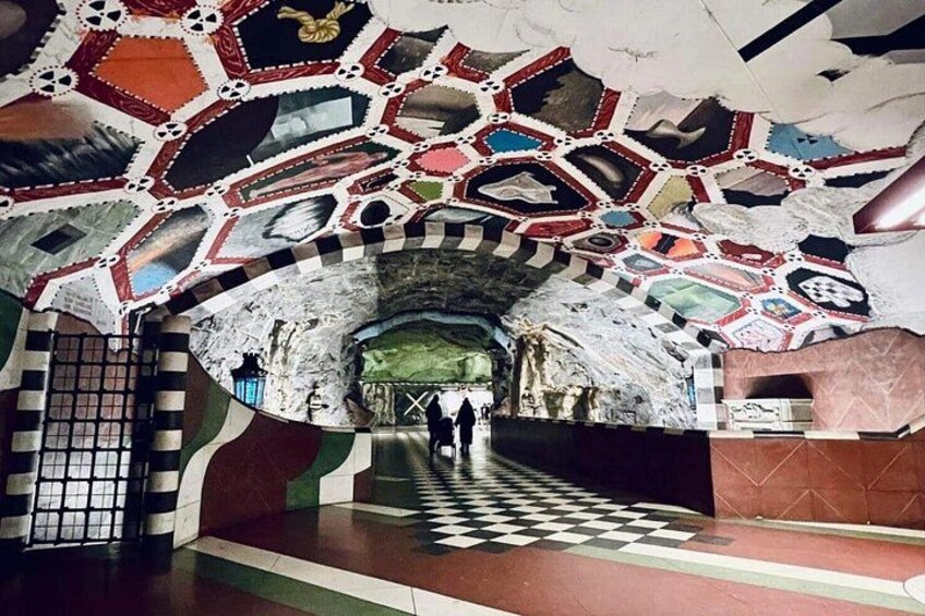 Une station de métro avec un sol à carreaux, une entrée de tunnel et un plafond orné d’une mosaïque colorée. 4
