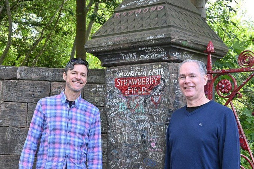 Dos hombres de pie frente a un muro con grafitis y un letrero que dice 'Strawberry Field'. 5