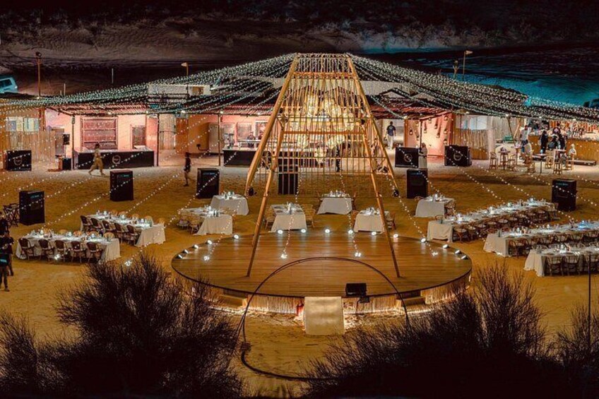 An outdoor event with tables arranged in a semi-circle, a central stage, and string lights. 1