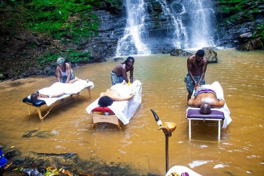 A natural setting with a waterfall, people receiving a massage in a river, and wooden massage tables. 2