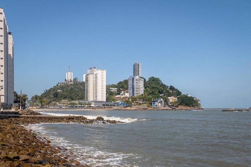 A coastal cityscape with a rocky shoreline, a clear blue sky, and several high-rise buildings on a hill. 2
