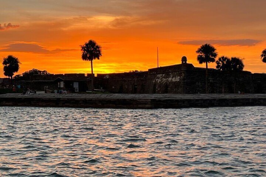 A sunset over a body of water with palm trees and a stone wall. 2