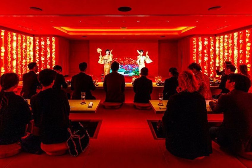 A dimly lit room with red ambient lighting, a stage with a performer, and an audience seated at tables. 5