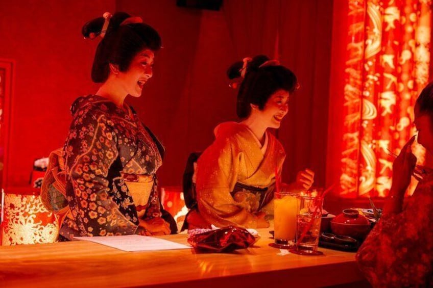 Two individuals in traditional attire at a table with a drink and a decorative backdrop. 1