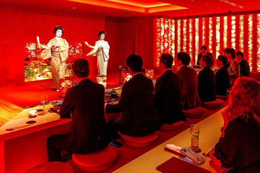 A traditional Japanese performance with two actors in kimonos, an audience seated at tables, and a stage with a red backdrop. 4