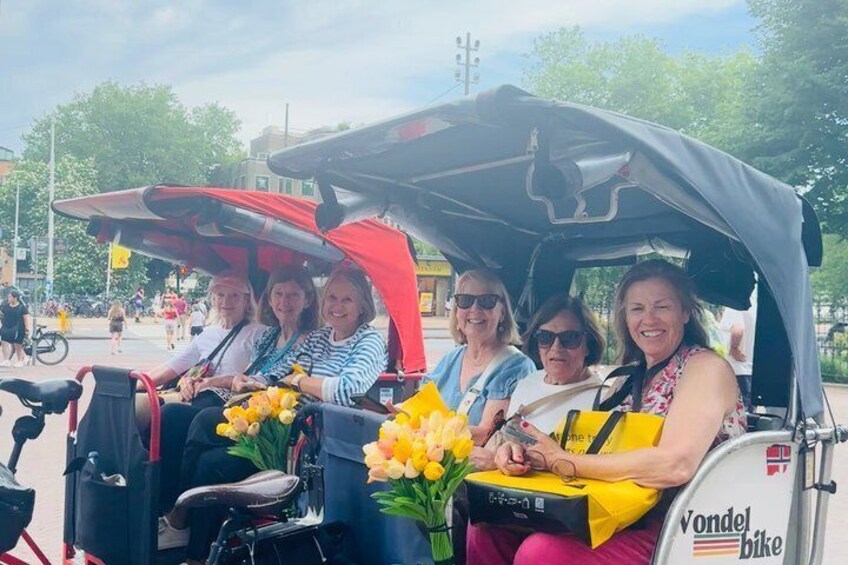 A group of people in a covered bicycle cart with flowers, parked on a street. 2
