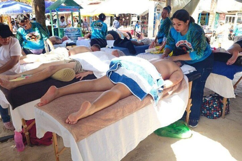 A massage parlor with clients lying on massage tables, a woman attending to a client, and a crowd of people in the background. 2
