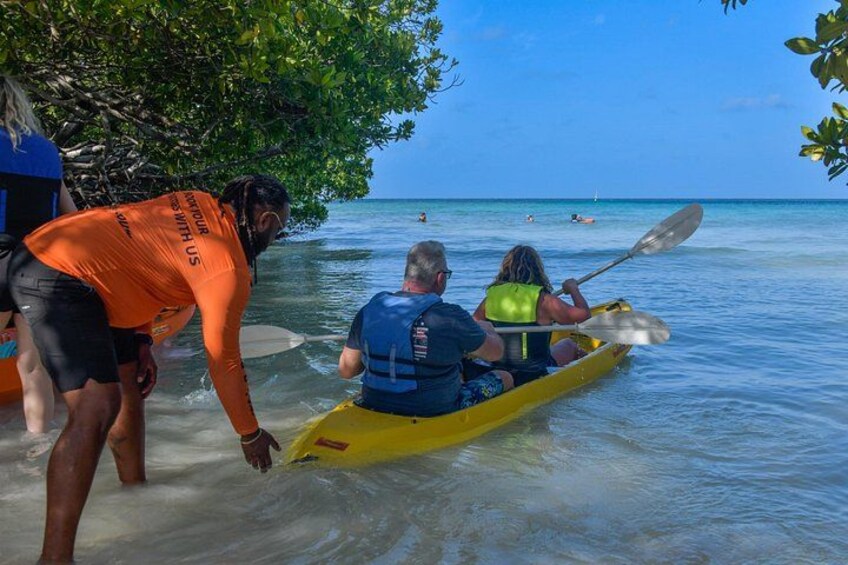 A person in a yellow kayak on calm water, another person in a blue life jacket, and a third person in an orange shirt assisting. 1