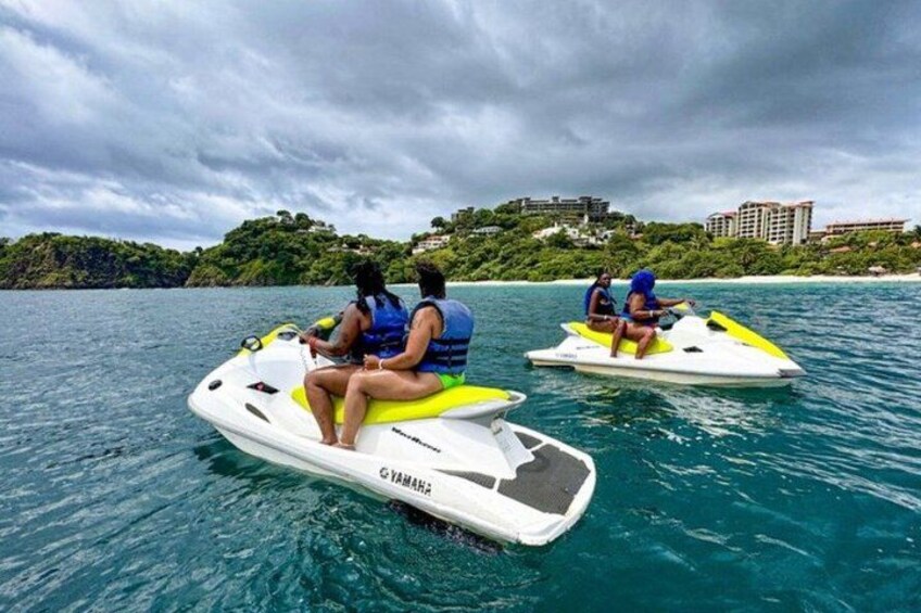 Two people jet skiing on a body of water with a cloudy sky and a distant shoreline with buildings. 4