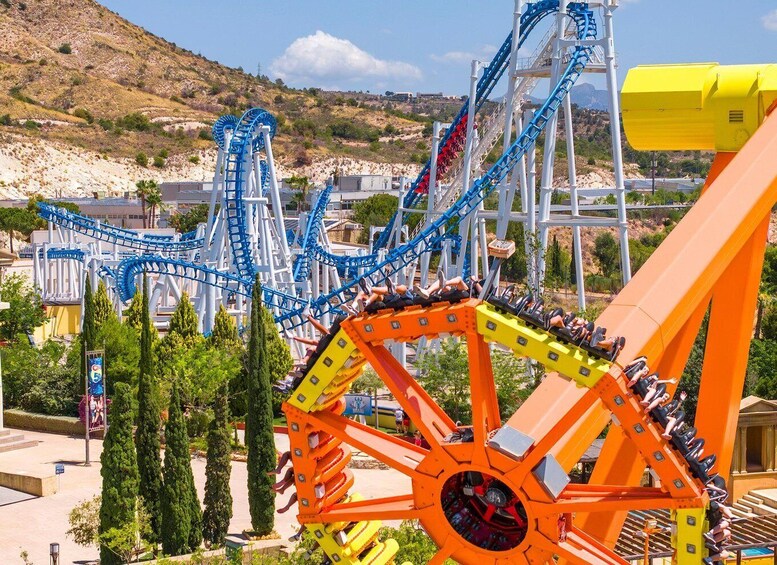 A roller coaster with blue and orange tracks, set against a hilly landscape with trees and buildings. 1