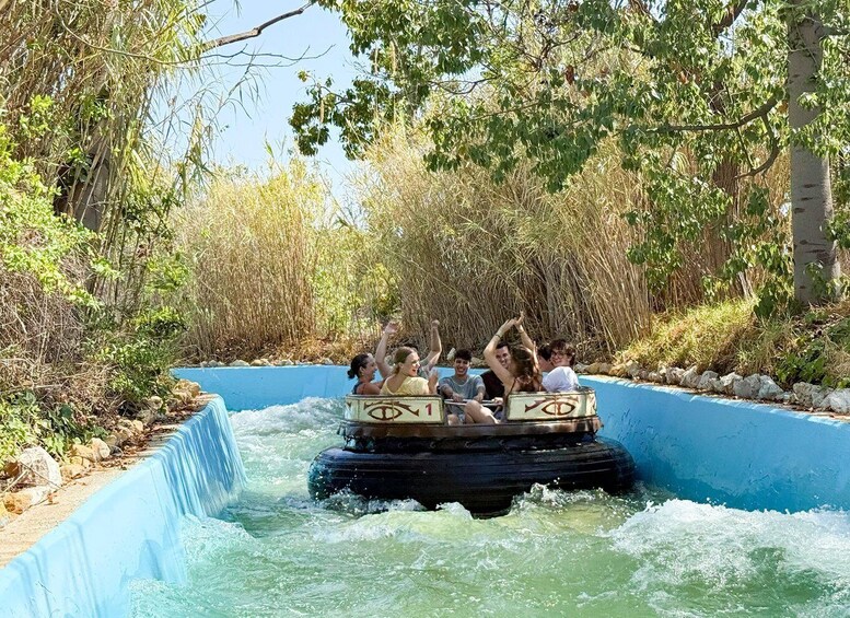People riding on a water ride with a blue channel and trees around. 3