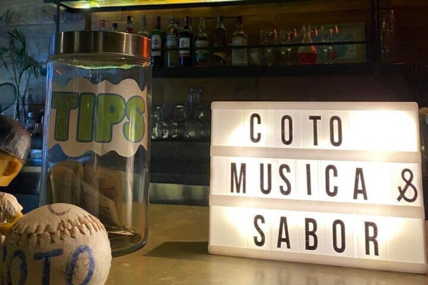 A bar counter with a light-up sign that reads "COTO MUSICA & SABOR" and a glass labeled "TIPS". 5