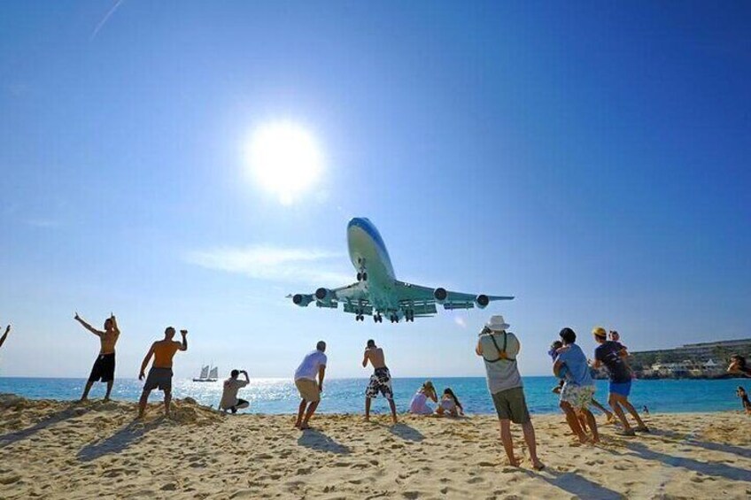 Un grande aereo vola a bassa quota sopra una spiaggia, mentre persone lo osservano. 1