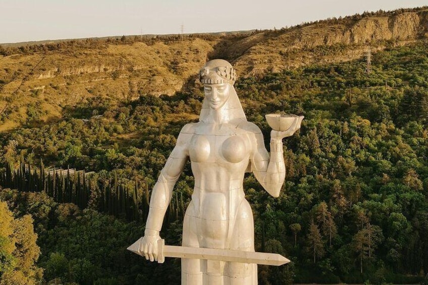 A large statue of a female figure holding a bowl and a spear, set against a mountainous backdrop. 1