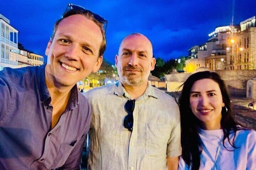Three people standing outdoors at dusk with buildings in the background. 2