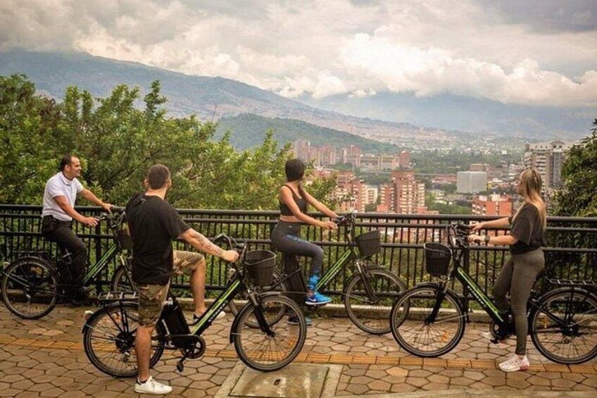 Eine Gruppe von Radfahrern, ein asphaltierter Weg und eine Stadtlandschaft mit Gebäuden und Bergen im Hintergrund. 1