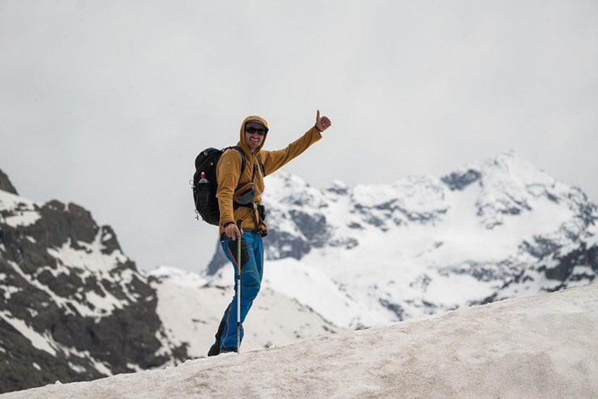A hiker in a yellow jacket and blue pants standing on a snow-covered slope with a mountain range in the background. 4