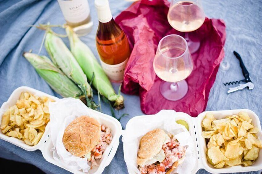 A picnic setup with sandwiches, chips, corn on the cob, wine glasses, and a bottle. 1