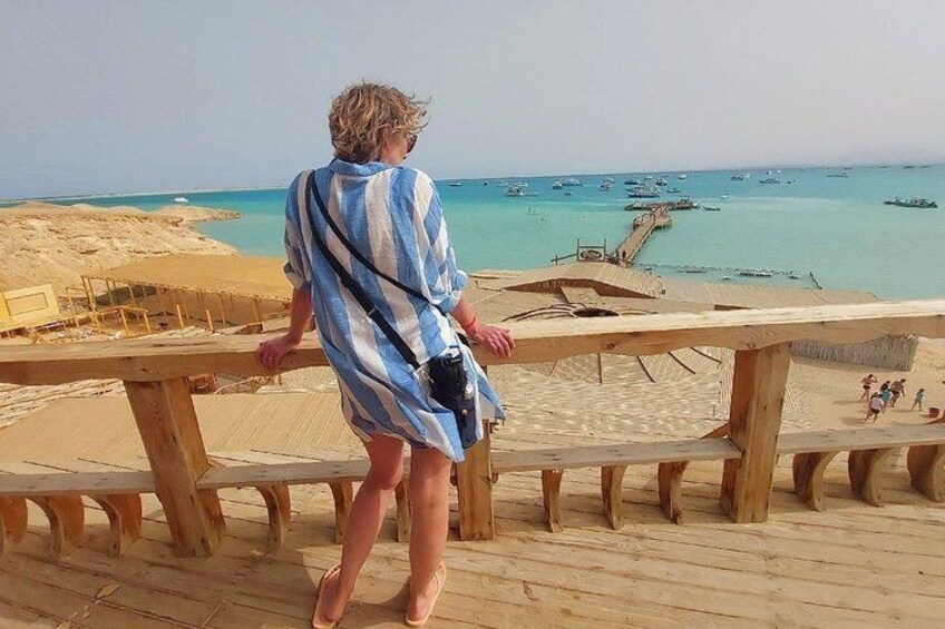 En person står på en træterrasse med udsigt over en strand, både og en mole. 4