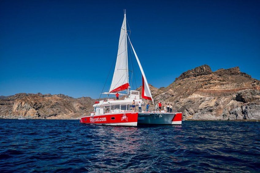 A red and white catamaran with the website 'obycat.com' on its side, sailing on calm waters with a rocky coastline in the background. 3