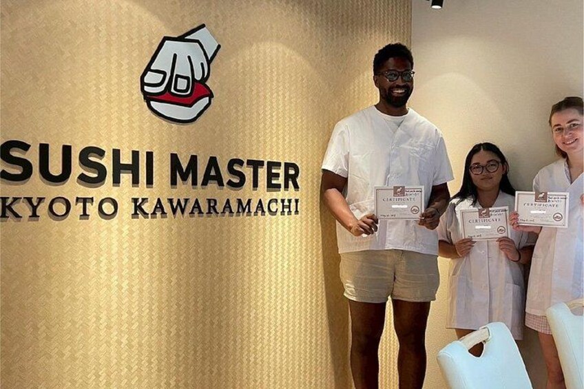 Tre personer holder certifikater foran en væg med teksten 'SUSHI MASTER KYOTO KAWARAMACHI'. 2