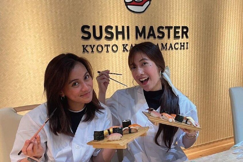 Dos personas con batas blancas sostienen bandejas de sushi frente a un letrero que dice 'SUSHI MASTER KYOTO KAMACHI'. 2