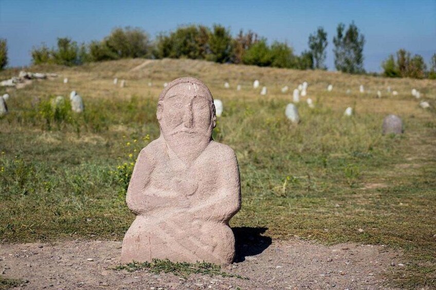 A stone statue of a seated figure in a grassy field with scattered gravestones in the background. 2