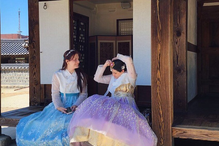 Two individuals in traditional Korean hanbok dresses sitting on a wooden bench outside a building with a traditional Korean roof. 2