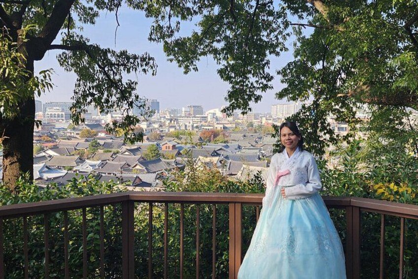 A person in traditional attire standing on a balcony with a cityscape in the background. 5