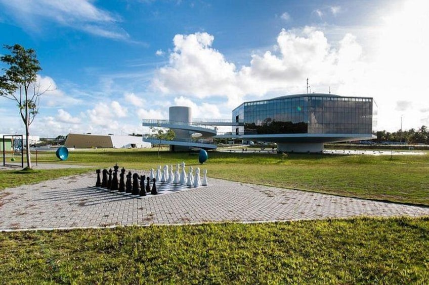 A chessboard on a paved area with a modern building and an airplane in the background. 4