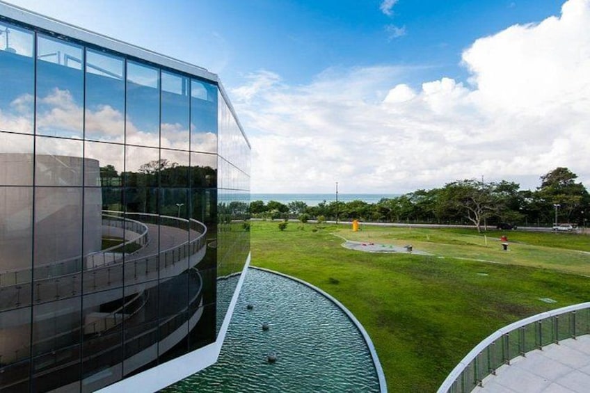 A modern building with a glass facade, a green park with a water feature, and a clear sky. 2