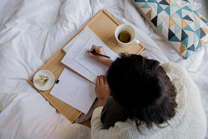 A person writing on a clipboard on a bed with a cup of tea and a geometric pillow. 2