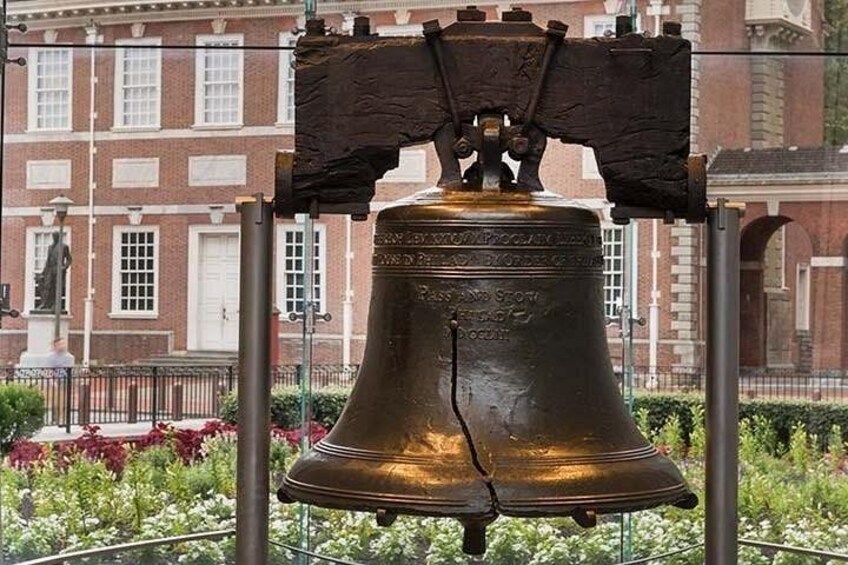 Eine große bronzene Glocke, die auf einem Podest mit einer Gedenktafel angebracht ist, vor einem Backsteingebäude mit Fenstern und einer Statue. 1