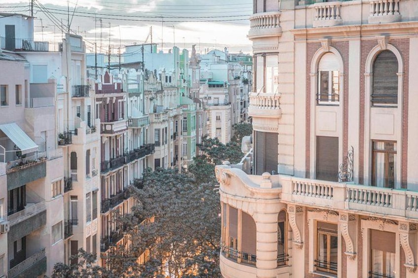 A densely packed urban street with multi-story buildings, balconies, and a tree-lined street. 2