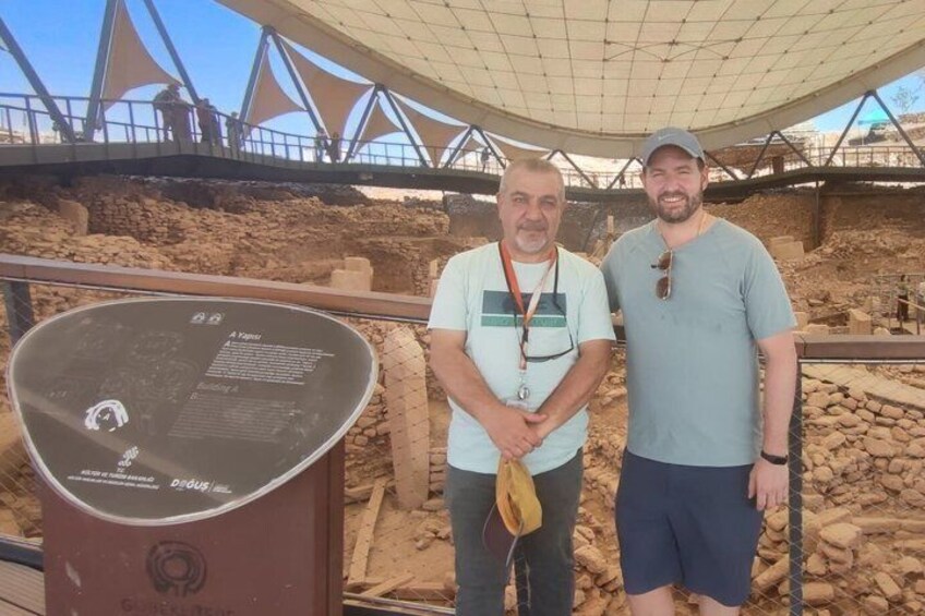 Two men standing in front of an archaeological site with a large informational display. 2