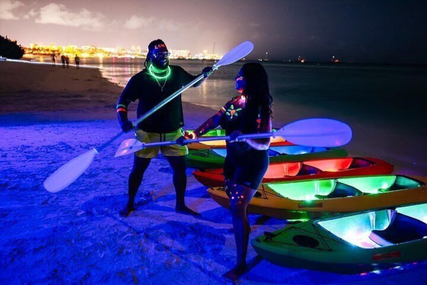 Two people standing on a beach with illuminated kayaks and a paddleboard. 2