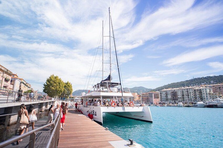 Ein Yachthafen mit einer weißen Yacht an Land, Personen auf einer Holzsteganlage und Gebäuden im Hintergrund. 2