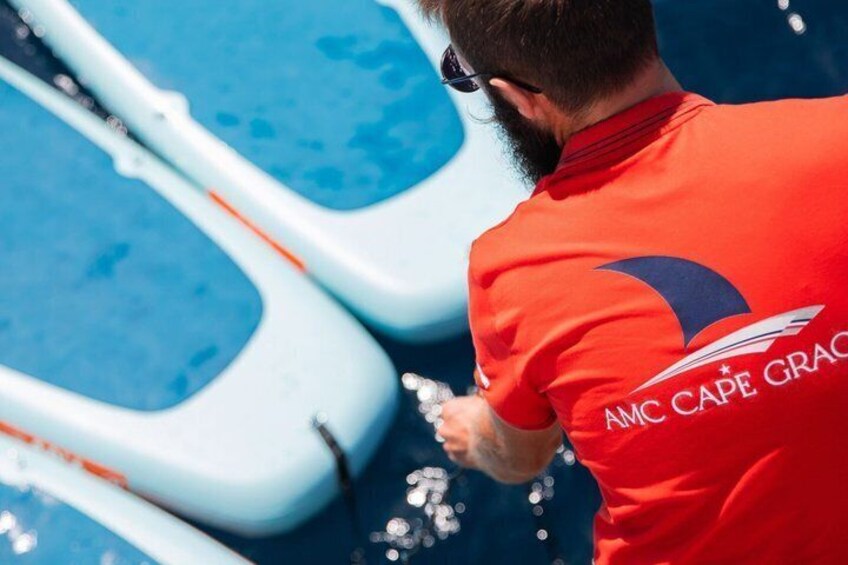 Una persona con una camiseta roja con el logotipo 'AMC Cape Grace' manejando un bote inflable blanco sobre una superficie de agua azul. 4