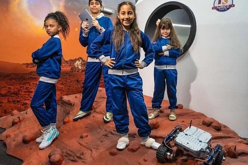 Quatre enfants, vêtus de combinaisons bleues sur le thème de l’espace, se tiennent sur une surface rouge, devant un robot et un décor sur le thème de l’espace. 3