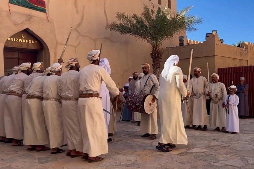A group of men in traditional attire standing in a line, some holding sticks, in front of a building with a palm tree. 3