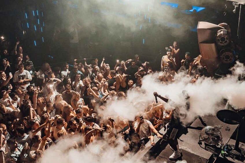 A crowded indoor event with people, a stage, and smoke effects. 2