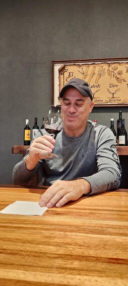 A person holding a wine glass, seated at a wooden table with bottles of wine in the background. 4