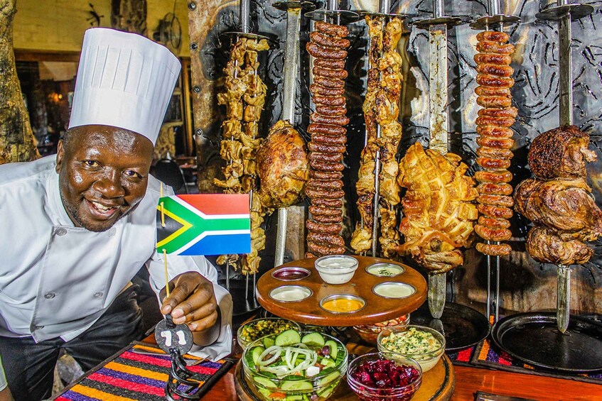 Uno chef tiene una bandiera sudafricana, con diversi spiedini di carne e insalate e salse in ciotole davanti a lui. 2