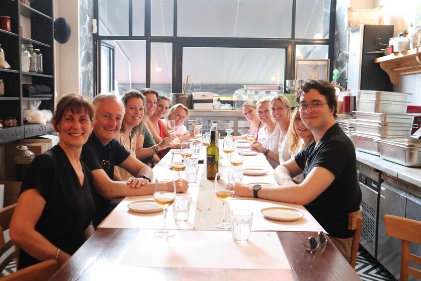 A group of people seated around a long dining table with wine glasses and bottles, in a room with shelves and kitchen equipment. 5