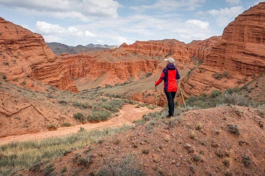 A hiker in a red jacket walking on a dirt trail in a red rock canyon. 1