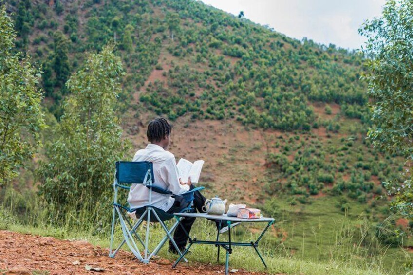 A person sitting on a chair in a grassy area with a book, a teapot, and a box on a small table. 3