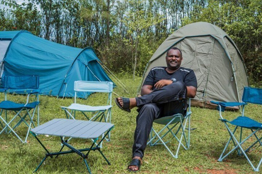 A person sitting on a camping chair in front of a tent, with another tent and chairs in the background. 1