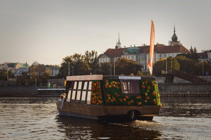 Een houten boot met een bloemenversierde cabine, drijvend op een rivier met een stadsgezicht op de achtergrond. 4