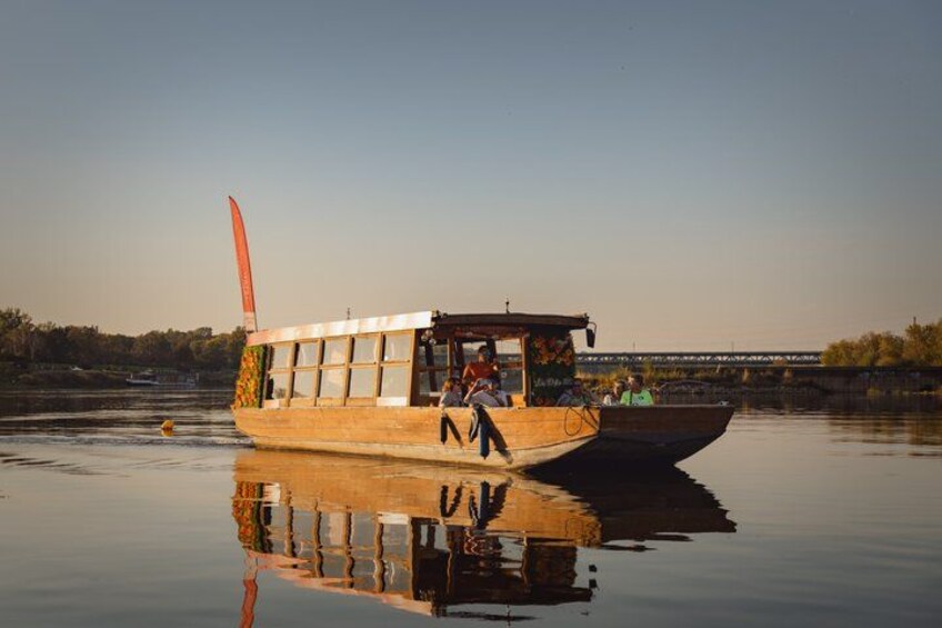 Een houten boot met passagiers, een brug op de achtergrond en rustig water dat het tafereel weerspiegelt. 3