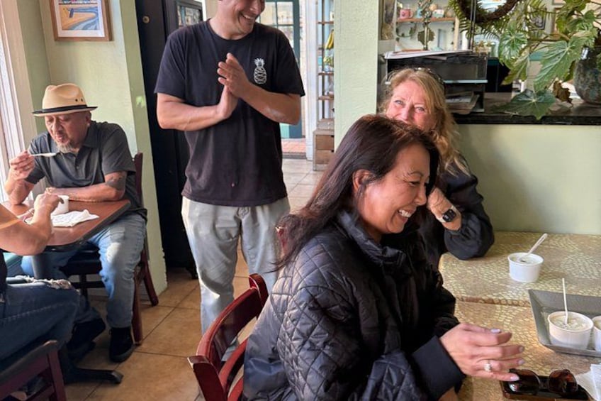 Un grupo de personas sentadas en una mesa en un café, con un hombre de pie aplaudiendo. 2